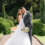 Portrait of happy married couple, handsome groom in stylish gray suit hugging bride in wedding dress with bouquet while posing in the garden. Wedding day ceremony.